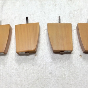 May include: Four wooden furniture legs with a light brown finish. Each leg has a rectangular body with a curved side and a metal rod extending from the top. The legs are designed for furniture support and stability.