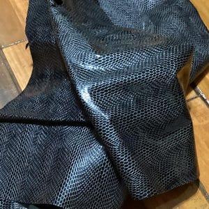 May include: Close-up of a folded piece of black and silver faux snakeskin fabric. The material has a textured, scale-like pattern and a glossy finish. The fabric is draped over a wooden surface, showcasing its flexibility and sheen.