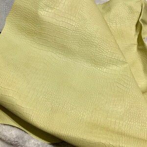 May include: A sheet of pale yellow leather with a crocodile skin texture. The leather is folded over, revealing the underside. The surface has a subtle sheen, and the texture is detailed.
