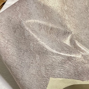 May include: A close-up of a light gray snakeskin fabric with a subtle shimmer. The fabric has a textured surface and a slightly wrinkled appearance.