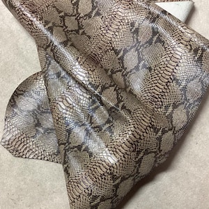 May include: A piece of faux snakeskin fabric with a detailed pattern in shades of brown, tan, and black. The material is folded, revealing the underside, which appears to be a lighter color. The fabric has a shiny, textured surface.