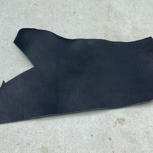 May include: A single, irregularly shaped piece of dark navy blue leather with a visible pebbled texture. The material features a smooth, matte finish and distinct, uneven edges, showcasing the natural grain and cut of the hide.