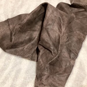 May include: A piece of brown leather fabric, folded and draped. The leather has a textured surface and a mottled appearance, with variations in color. The edges of the leather are visible, and the fabric is resting on a white, fuzzy surface.