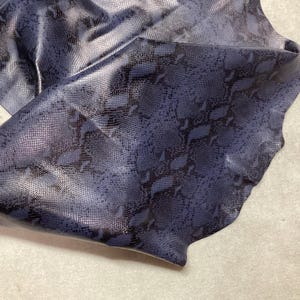 May include: A piece of blue and black snakeskin leather. The leather has a shiny, textured surface with a scale pattern. The material is folded over, revealing the underside. The background is a light beige color.