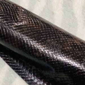 May include: Close-up of a rolled piece of dark brown leather with a snakeskin pattern. The leather has a glossy finish and a textured surface, with a grid-like design. The material is likely used for crafting.