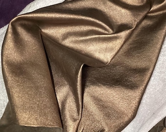 Könnte beinhalten: Nahaufnahme eines gefalteten Stücks schimmernden, bronzefarbenen Stoffes. Das Material hat einen metallischen Glanz und scheint weich und geschmeidig zu sein, mit sichtbaren Falten und Knitterstellen. Der Stoff ist über einer weißen Oberfläche drapiert.
