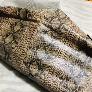 May include: Close-up of a rolled piece of faux snakeskin fabric. The fabric features a detailed pattern in shades of brown, tan, and gray, mimicking the texture and coloration of snake scales. The material appears to be flexible and possibly used for crafting.