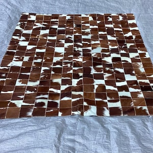 May include: A rectangular area rug constructed from a patchwork of square cowhide tiles. The rug features a repeating pattern of brown, white, and dark brown fur patches, creating a multi-tonal, textured surface with visible stitching.