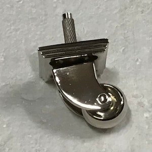 May include: A silver metal furniture caster with a square base and a threaded stem. The caster has a single wheel with a smooth, rounded surface.