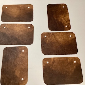 May include: Six brown leather patches with two holes each. The patches are rectangular and have a slightly textured surface.