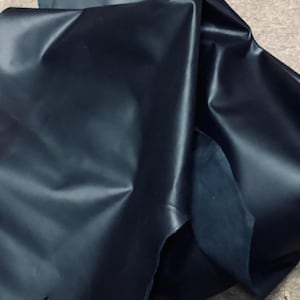 May include: Dark navy blue leather fabric, with a smooth, shiny surface. The material is folded and draped, showing the texture and sheen of the leather. A small hole is visible in the fabric.