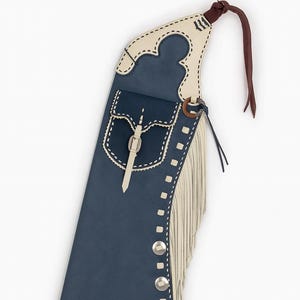 May include: A navy blue leather chaps with cream-colored accents. Features include a pocket, fringe, and decorative stitching. Silver-colored buttons and a leather strap with a buckle are also visible. A brown leather strap is attached at the top.