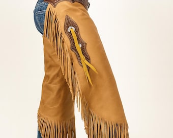 May include: Tan leather chaps with long fringe detail. The chaps have a decorative leather patch with a silver grommet and yellow leather accents. The chaps are worn over blue jeans and brown cowboy boots.