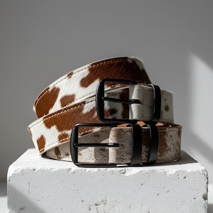 Puede incluir: Dos cinturones de piel de vaca con hebillas de metal negro apilados sobre una superficie blanca. Los cinturones tienen un patr&oacute;n moteado marr&oacute;n y blanco. Las hebillas son rectangulares y tienen un acabado mate. Los cinturones son de cuero.