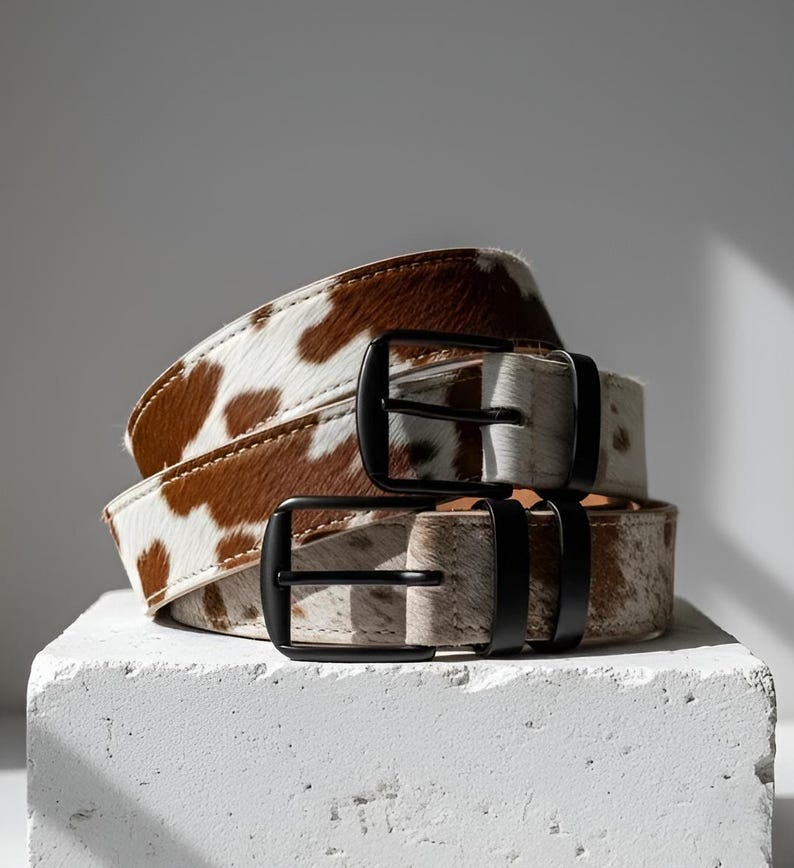 Puede incluir: Dos cinturones de piel de vaca con hebillas de metal negro apilados sobre una superficie blanca. Los cinturones tienen un patr&oacute;n moteado marr&oacute;n y blanco. Las hebillas son rectangulares y tienen un acabado mate. Los cinturones son de cuero.