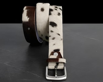 May include: A stylish belt with a cowhide pattern. The belt features a silver-colored buckle and a brown leather backing. The cowhide has a white base with brown spots. The belt is designed to be worn around the waist.