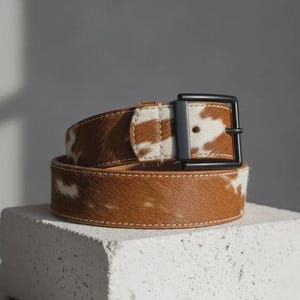 Puede incluir: Un cintur&oacute;n de piel de vaca marr&oacute;n y blanco con una hebilla de metal negro. El cintur&oacute;n est&aacute; hecho de cuero genuino y presenta un borde cosido. El cintur&oacute;n est&aacute; enrollado y se muestra sobre una superficie blanca.