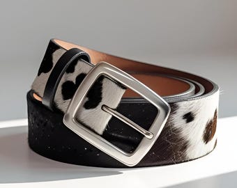 Puede incluir: Un cinturón elegante con un estampado de piel de vaca en blanco y negro. El cinturón presenta una hebilla cuadrada plateada y una correa de cuero negro. El ancho del cinturón es de aproximadamente 3,8 cm.