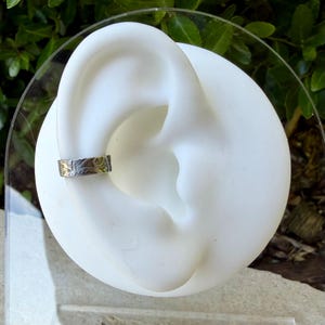 May include: A silver ear cuff with a detailed floral design. The cuff is displayed on a white ear-shaped form, showcasing the jewelry's intricate pattern. The background features green foliage.
