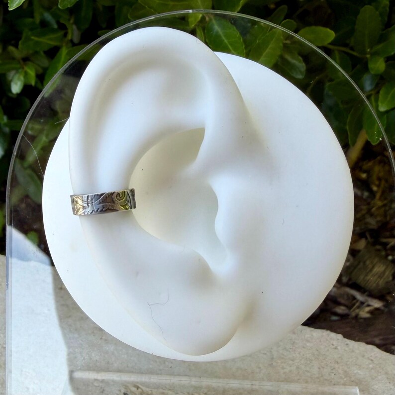 May include: A silver ear cuff with a detailed floral design. The cuff is displayed on a white ear-shaped form, showcasing the jewelry's intricate pattern. The background features green foliage.