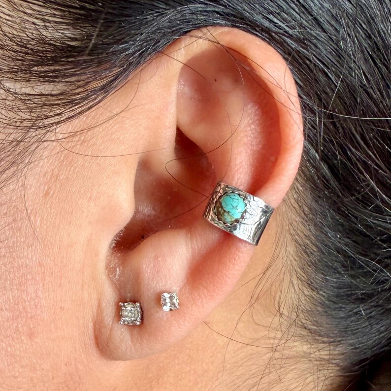 May include: A silver ear cuff with a turquoise stone and a floral design is worn on an ear. Two small diamond stud earrings are also visible. The ear cuff is a unique piece of jewelry.