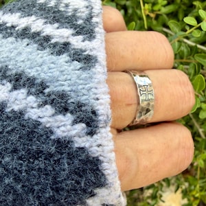 May include: A silver-toned metal ring with a hammered, textured surface and a prominent cross symbol engraved on the front. The band has a wide, flat profile and a polished finish, showcasing a distinct cross design centered on the outer surface.