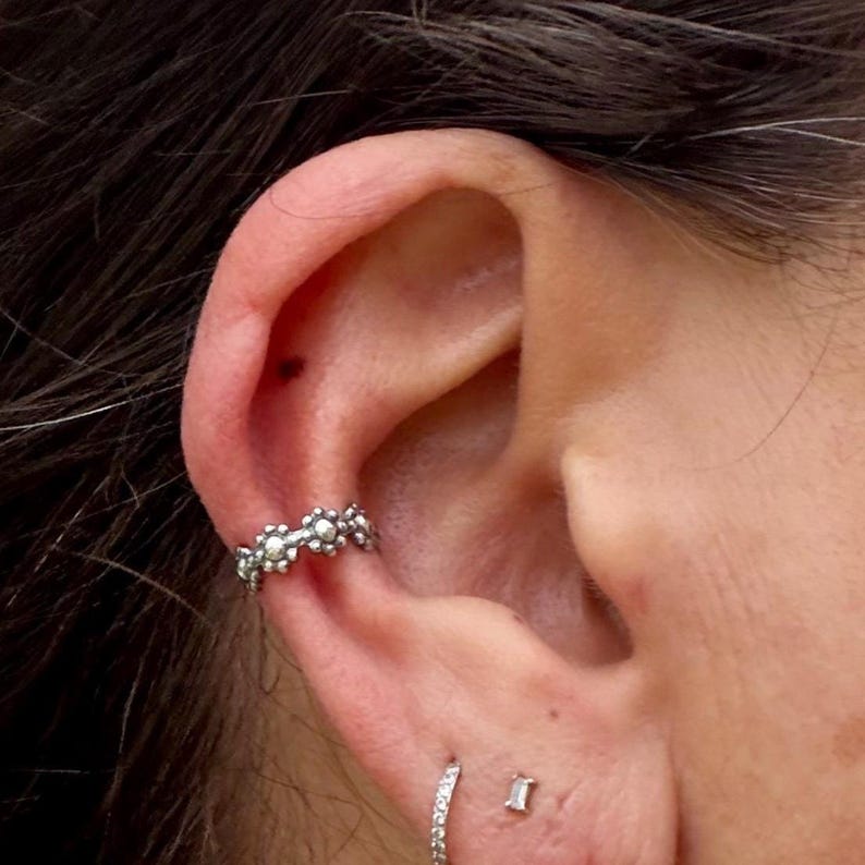 Puede incluir: Un brazalete de oreja plateado con un dise&ntilde;o floral se usa en la parte superior de la oreja. Tambi&eacute;n se ven un peque&ntilde;o pendiente de aro y un pendiente de bot&oacute;n. La oreja est&aacute; colocada sobre un fondo de cabello oscuro.