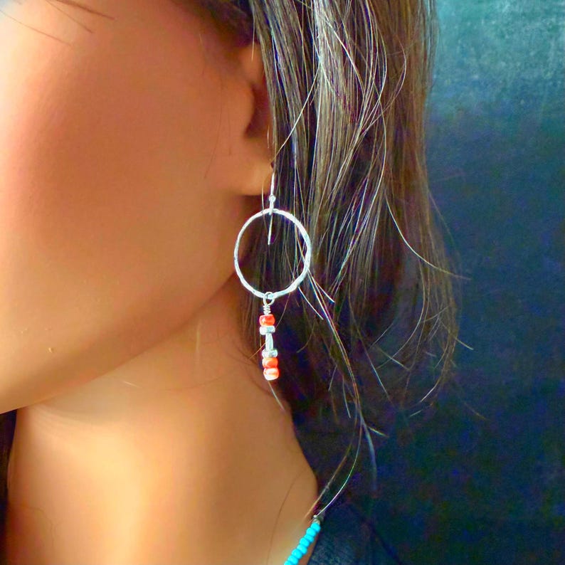 May include: A silver-toned dangle earring featuring a thin circular hoop wire. Suspended from the bottom of the hoop is a vertical arrangement of small, cylindrical beads in alternating shades of bright orange and metallic silver.