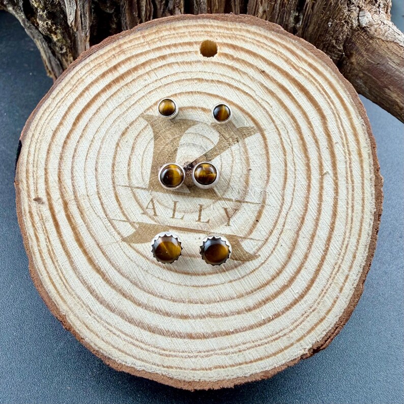 Puede incluir: Tres pares de pendientes de bot&oacute;n con piedras preciosas de ojo de tigre redondas y pulidas engastadas en metal plateado. El par superior es peque&ntilde;o y liso, el par central es de tama&ntilde;o mediano y el inferior tiene un borde dentado.
