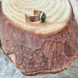 May include: A gold-colored adjustable ring with a turquoise stone. The ring's band features an etched floral design. The turquoise stone is oval-shaped with brown and blue hues. The ring is displayed on a wooden surface.
