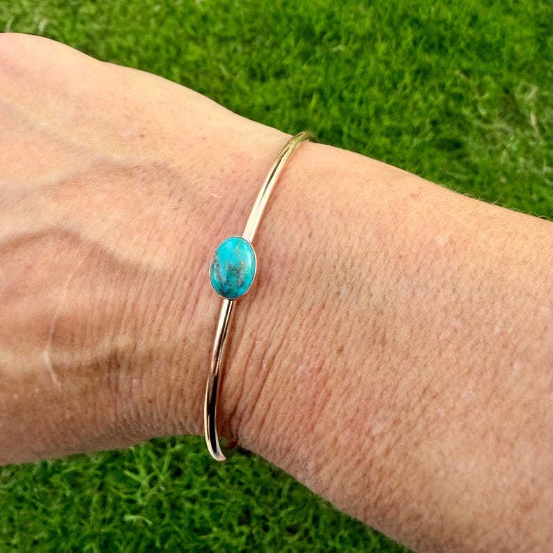May include: A gold-colored bangle bracelet with a turquoise stone. The oval-shaped turquoise stone is set in the center of the bracelet. The bracelet is on a person's wrist, with a green background.