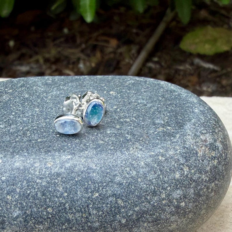 May include: A pair of silver stud earrings with oval-shaped, light blue and teal gemstones. The earrings are set on a gray stone surface, with a blurred green background.