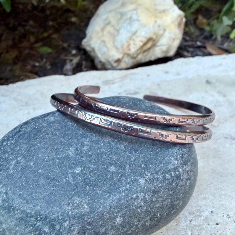 May include: Two copper cuff bracelets featuring a repeating geometric pattern engraved along the outer surface. The bracelets have an open-ended design and a polished metallic finish, showcasing a warm reddish-brown colour with dark recessed details.