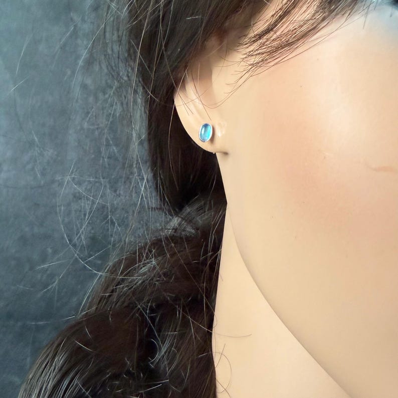 May include: A close-up of a person's ear with a small, oval-shaped, light blue stud earring. The earring has a silver or white metal setting. The background is a dark, textured surface.