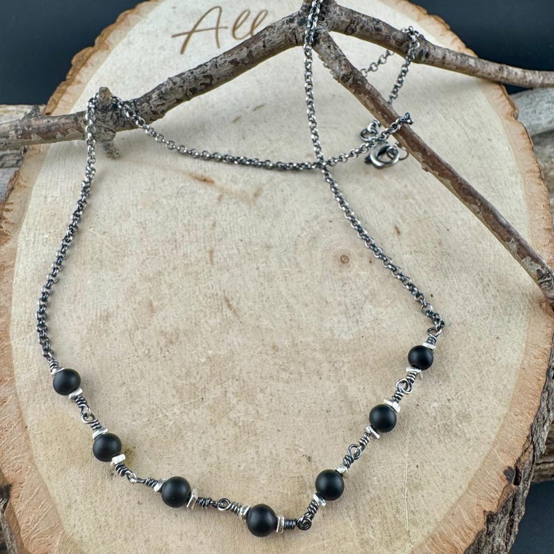 May include: A silver-toned necklace with black beads. The necklace features a delicate chain and is adorned with several round, matte black beads. The beads are spaced along the chain and are secured with silver wire. The necklace is displayed on a wooden surface.