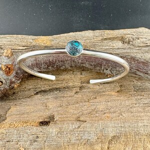 May include: A silver cuff bracelet with a round, turquoise-colored stone set in the center. The bracelet has a simple, open design and is displayed on a piece of wood.