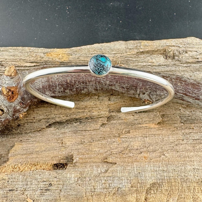 May include: A silver cuff bracelet with a round, turquoise-colored stone set in the center. The bracelet has a simple, open design and is displayed on a piece of wood.