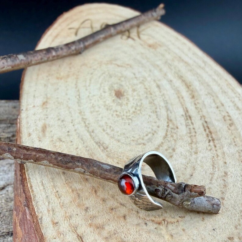 May include: A silver ring with a textured band and a round, orange-red gemstone. The ring is displayed on a small wooden branch, set against a light brown wooden background.