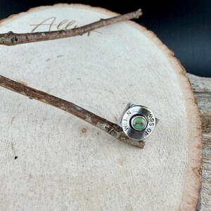 May include: A silver ring featuring a turquoise stone set within a bullet casing. The casing is engraved with the text "WLL N S&W 40". The ring is presented on a wooden surface, with a twig resting nearby.