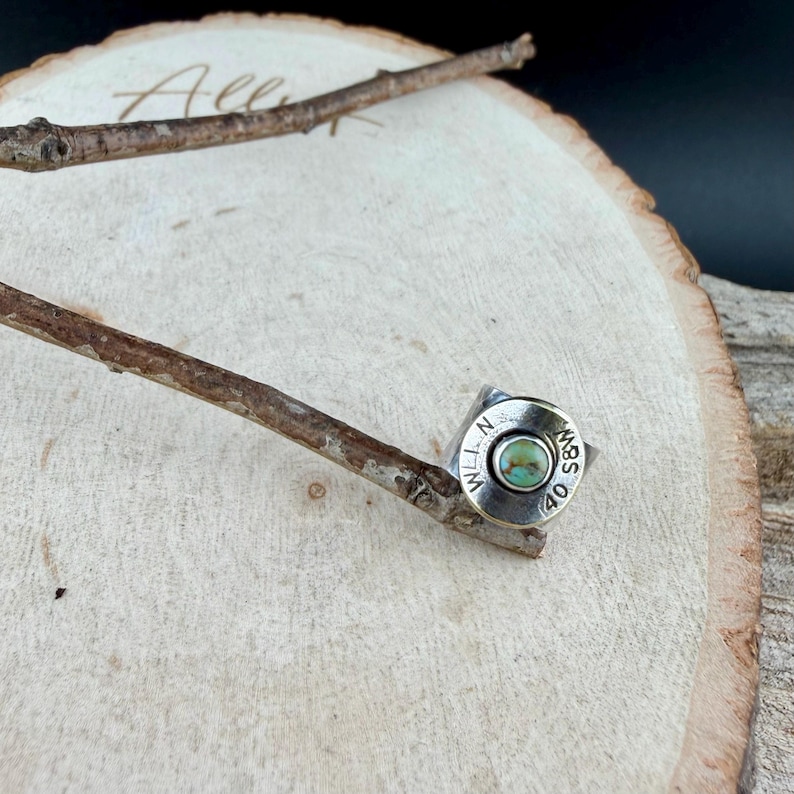 May include: A silver ring featuring a turquoise stone set within a bullet casing. The casing is engraved with the text "WLL N S&W 40". The ring is presented on a wooden surface, with a twig resting nearby.