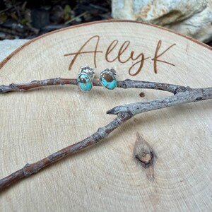 May include: A pair of oval turquoise stud earrings with silver settings. The earrings are displayed on a wooden slice with the name "Allyk" engraved on it. The earrings are set on a small branch.