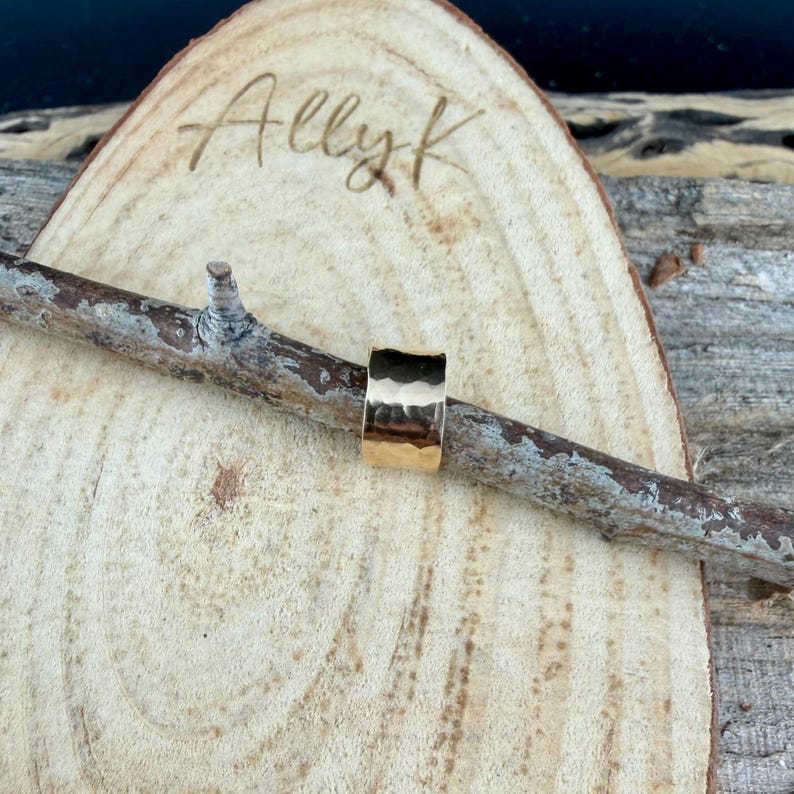 Puede incluir: Un anillo dorado con textura martillada. El anillo se muestra sobre una peque&ntilde;a rama, sobre un fondo de madera. El texto "Ally K" es visible en la superficie de madera.