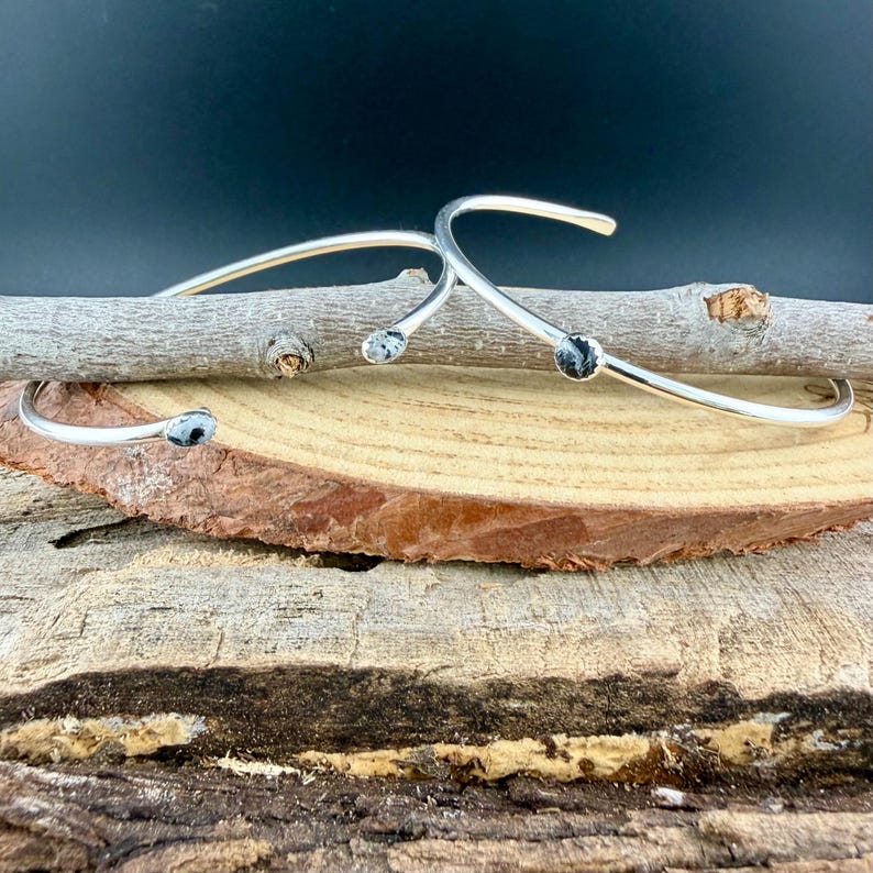 May include: Three silver-toned, open cuff bracelets with a curved design. Each bracelet features a small, round, black and white speckled stone at the ends. The bracelets are displayed on a wooden surface with a dark background.