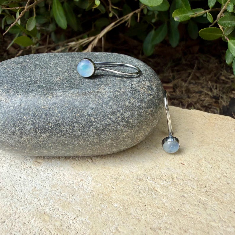 May include: A pair of silver-toned earrings featuring a single round, iridescent cabochon stone set in a smooth metal frame. The earrings have a curved, open-loop design that extends from the stone setting, creating a sleek and simple silhouette.
