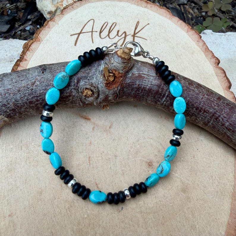 May include: A turquoise and black beaded bracelet with silver accents. The bracelet features oval turquoise stones interspersed with black beads and silver spacers. The bracelet is secured with a silver clasp. The name "Allyk" is written on the wooden surface.