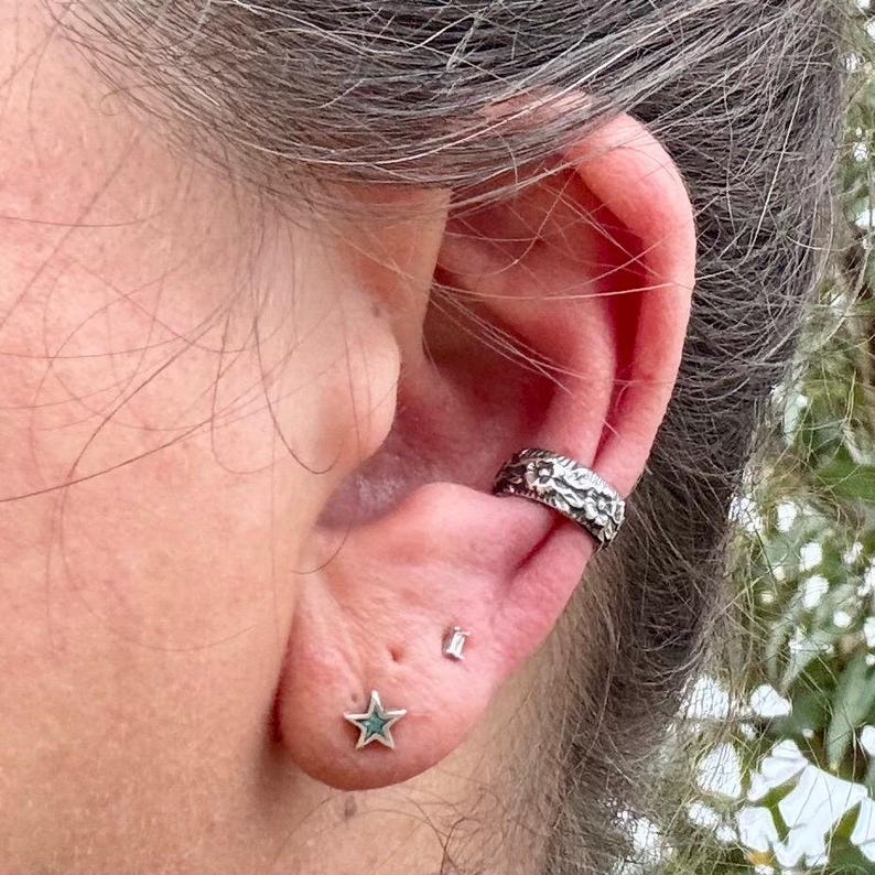 May include: Close-up of an ear adorned with jewelry. A silver ear cuff with a textured floral design is on the upper ear. A star-shaped stud earring with a turquoise center and a small rectangular stud are also visible.