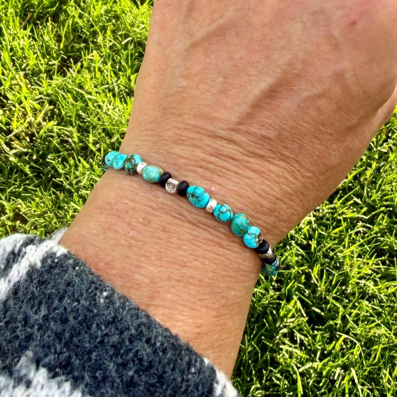 May include: A turquoise beaded bracelet with silver accents and black beads. The bracelet is strung on a stretchy cord and is worn on a wrist. The background is a blurred view of green grass.