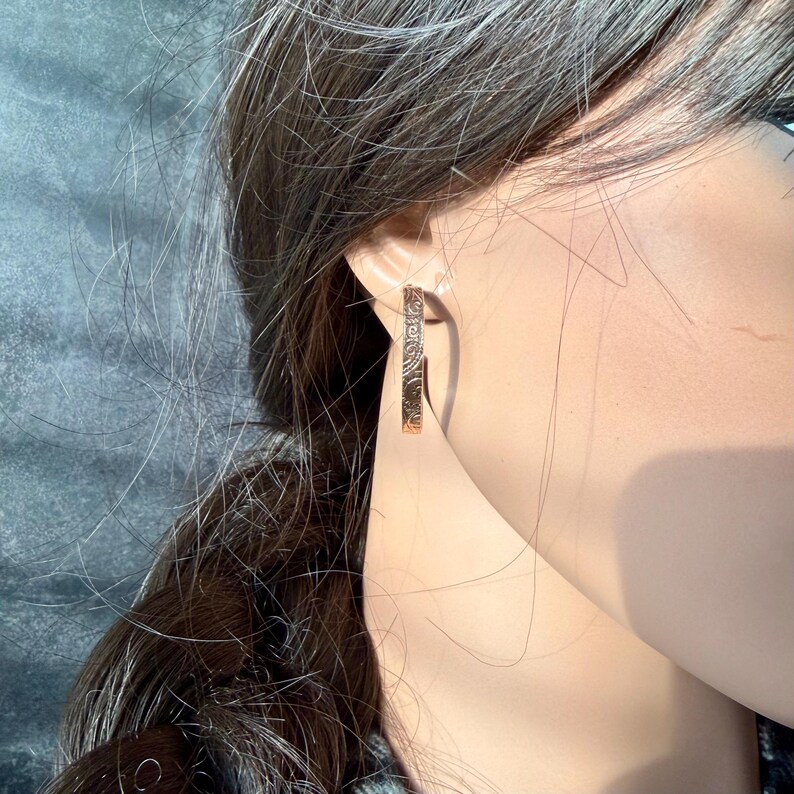 May include: Rose gold hoop earrings with an engraved floral pattern. The earrings are worn on a person's ear, with the design visible. The earrings are a warm, metallic color and have a simple, elegant design.