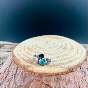 K&ouml;nnte beinhalten: Ein verstellbarer Ring in Silberoptik mit einem T&uuml;rkisstein. Der Ring hat ein offenes Design und ist mit ge&auml;tzten Mustern verziert. Der T&uuml;rkisstein ist rund und in einer L&uuml;nettenfassung gefasst. Der Ring wird auf einer Holzoberfl&auml;che pr&auml;sentiert.