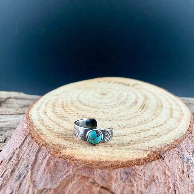 K&ouml;nnte beinhalten: Ein verstellbarer Ring in Silberoptik mit einem T&uuml;rkisstein. Der Ring hat ein offenes Design und ist mit ge&auml;tzten Mustern verziert. Der T&uuml;rkisstein ist rund und in einer L&uuml;nettenfassung gefasst. Der Ring wird auf einer Holzoberfl&auml;che pr&auml;sentiert.